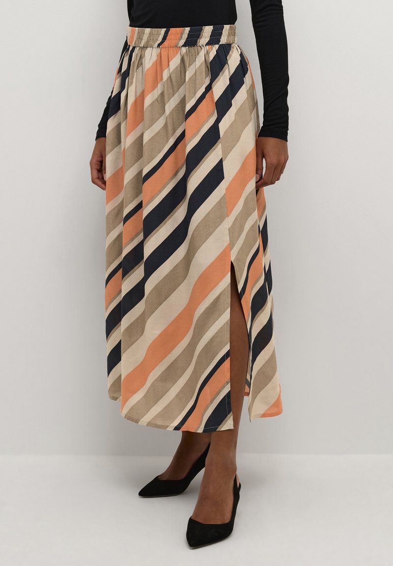 Femme portant une jupe midi rayée avec des rayures diagonales beige, noir et orange et une fente latérale, associée à des chaussures noires à bout pointu.