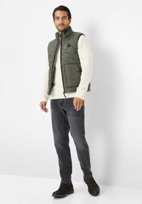Olivenfarbene Steppweste mit Reißverschluss und seitlichen Taschen. Trägt man über einem cremefarbenen Pullover und kombiniert mit dunklen Jeans und Stiefeln.