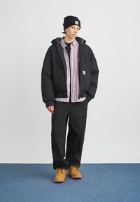 Melna izolēta jaka virs svītraina pogājama krekla, melnas bikses un brūnas zābaki. Cepure ir melna ar logotipu. Vienkāršs, funkcionāls dizains.