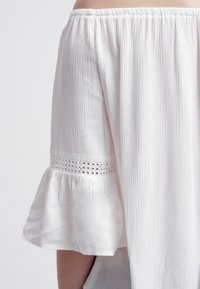 Vit off-shoulder topp med en texturerad tyg, volangärmar och spetsdetaljer vid ärmsluten. Lätt och svävande design.