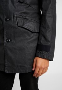 Parka nera, impermeabile, con molteplici tasche frontali, chiusure a bottoni e tessuto testurizzato. Presenta un dettaglio in elastico nero ai polsini.