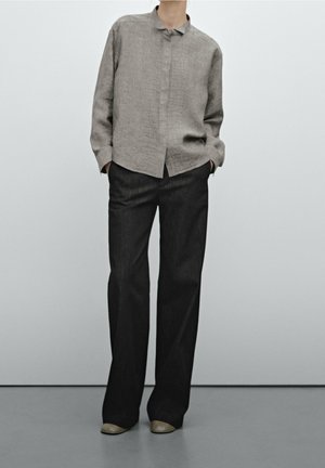 Osoba ubrana w szarą koszulę na guziki z długim rękawem, czarne spodnie o szerokich nogawkach oraz beżowe buty na jednolitym jasnoszarym tle.