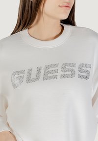 Biała bluza z okrągłym dekoltem, długimi rękawami i napisem "GUESS" z srebrnych cyrkonii na piersi. Miękka tekstura materiału.