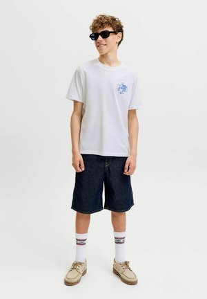 Ung person iført sorte solbriller, hvid grafisk t-shirt, mørke denimshorts, hvide mærkesokker og beige sko, stående på hvid baggrund.