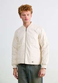 Lys beige bomberjakke med quiltet mønster, frontlynlås og sidelommer. Ribbet manchetter og kant, båret over blå denimjeans.