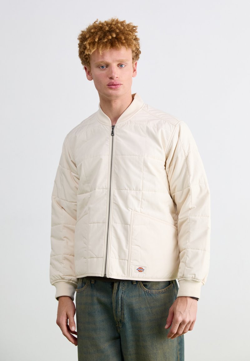 Lys beige bomberjakke med quiltet mønster, frontlynlås og sidelommer. Ribbet manchetter og kant, båret over blå denimjeans.