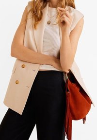 Gilet double boutonnage beige avec des boutons dorés, haut blanc côtelé, pantalon noir et sac en suède orange avec des franges.