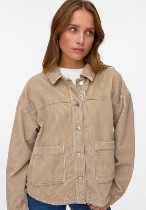 Jeune femme portant une veste beige en velours côtelé avec des boutons sur le devant et de grandes poches, regardant légèrement vers sa droite sur un fond blanc uni.
