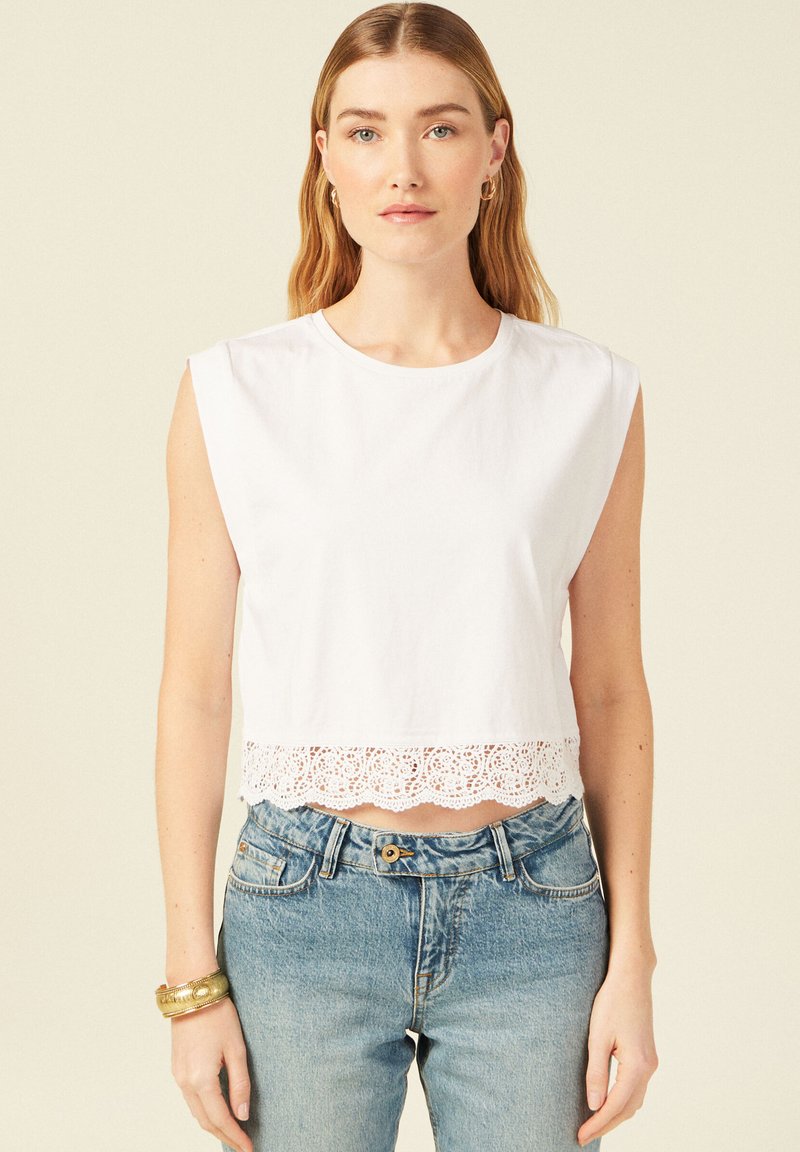 Witte mouwloze crop top van katoen, met een golvende kanten rand aan de onderzijde. Gecombineerd met lichtgewassen denim jeans.