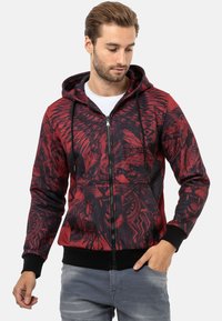 Roter Reißverschluss-Hoodie mit einem schwarzen, kunstvollen Grafikdesign, strukturiertem Stoff, Kordelzugkapuze und schwarzen Rippbündchen sowie -saum.