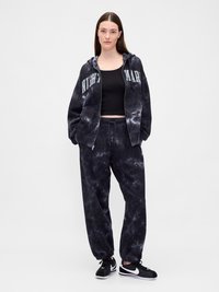 Γυναίκα φορώντας μαύρο φούτερ με tie-dye και ταιριαστά παντελόνια φόρμας, μαύρο crop top και άσπρα-μαύρα αθλητικά παπούτσια, standing με τα χέρια στις τσέπες.