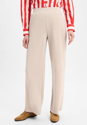 Beige weite Hosen aus weichem Stoff mit einem hohen Taillenschnitt. Kombiniert mit braunen Wildlederslippern und einem rot-weiß gestreiften Hemd.