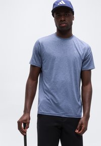 Άνδρας που φοράει μπλε καπέλο Adidas και μπλε ριγέ μπλουζάκι Calvin Klein, κρατώντας ένα μαύρο αντικείμενο με φόντο ένα απλό λευκό φόντο.