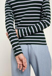Person in einem ribbed, langärmeligen dunkelgrün und weiß gestreiften Shirt, mit verschränkten Händen hinter dem Rücken, kombiniert mit hellblauen Hosen.