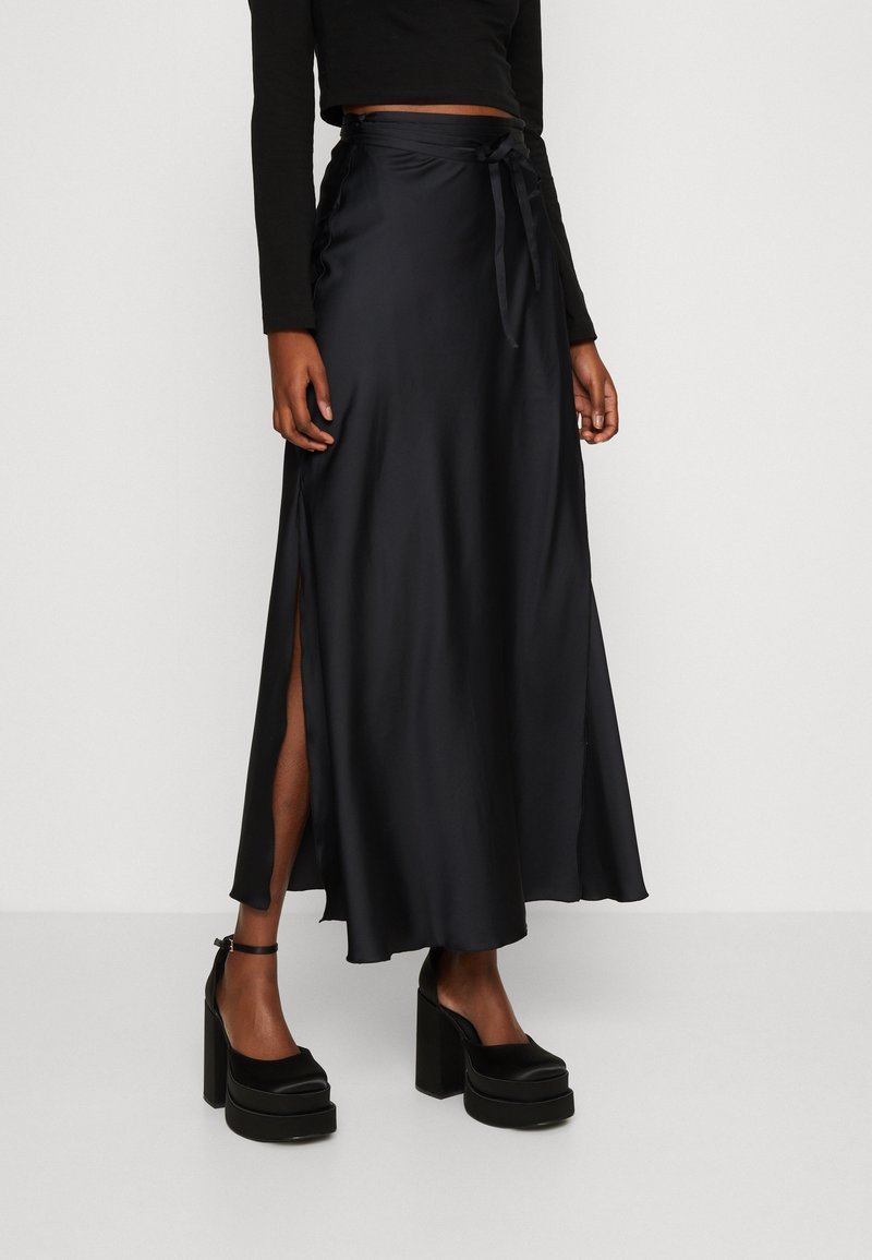Jupe maxi en satin noir avec fentes sur les côtés, taille à nouer et texture lisse. Associée à des chaussures à plateforme noires épaisses.