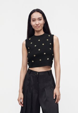 Jeune femme aux longs cheveux noirs porte un crop top noir sans manches avec des cercles dorés et un pantalon large noir taille haute.