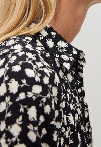 Schwarze Bluse mit weißem Blumenmuster, kragenförmiges Design, strukturiertes Material und Falten an den Schultern für zusätzliche Details.