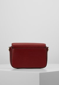 Borsa a tracolla compatta in pelle rossa scura con hardware in tonalità oro, texture granulosa e chiusura a pattina, posizionata su un pedestal bianco.