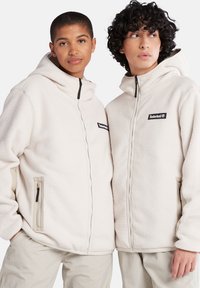 Cremfarbene Fleecejacken mit Reißverschluss und Kapuzen. Merkmale sind schwarze Timberland-Logos, Seitentaschen und strukturierter Stoff.