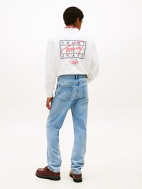 Sudadera blanca con un logo gráfico en la parte posterior, combinada con jeans holgados de un tono azul claro. Zapatos marrones con acabado texturizado completan el conjunto.