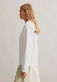 Witte sweatshirt met een ronde hals en geribde manchetten. Bevat blauwe tekst op de achterkant. Het model draagt een lichtgekleurde rok.