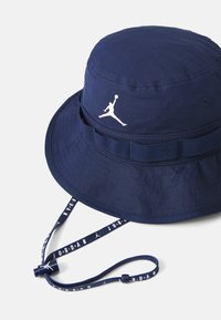 Navy blue bucket hat made of lightweight fabric, featuring a white Jumpman logo, adjustable chin strap, and textured band around the rim.