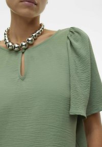 Top verde texturizzato con maniche a sbuffo e scollatura a chiave. Accentuato con una collana in argento con perle grandi.