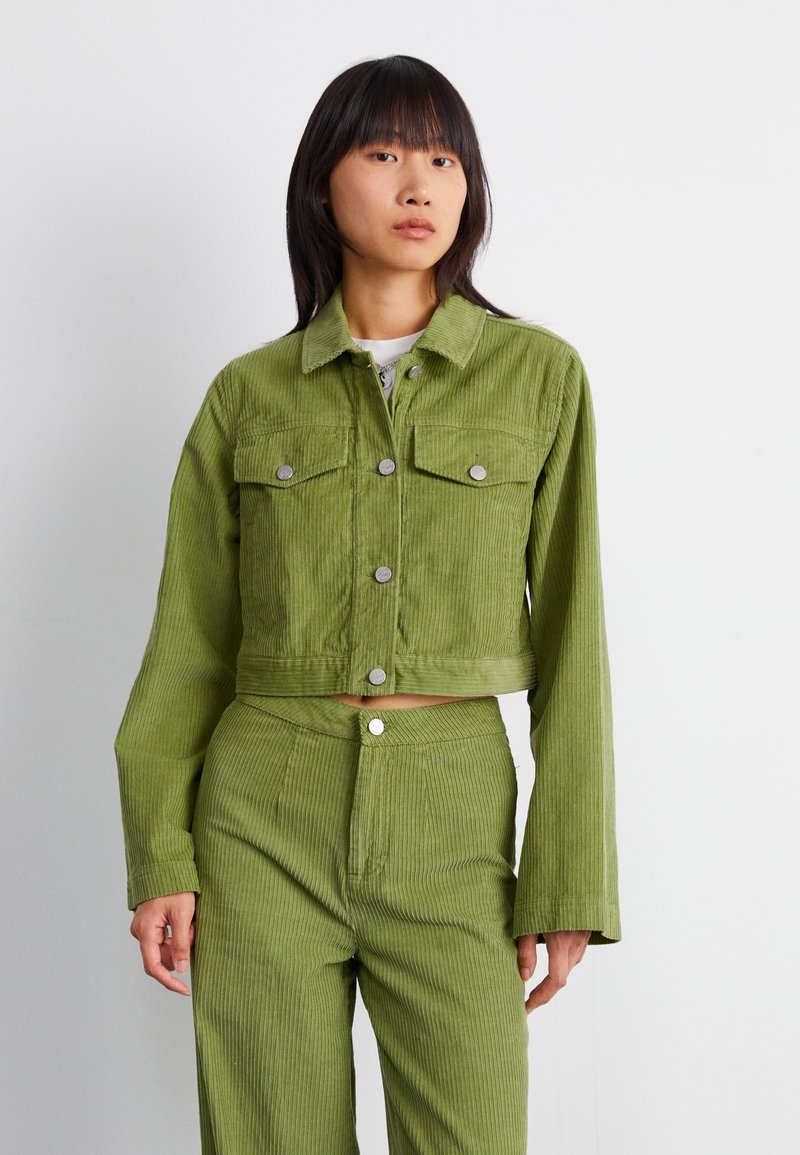 Chaqueta de pana verde recortada con dos bolsillos en el pecho, botones plateados y mangas largas. Combinada con pantalones de pana a juego.