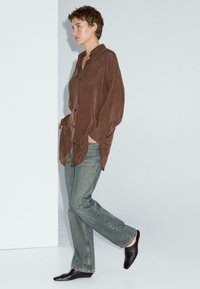 Persona che cammina indossando una camicia marrone chiara con bottoni, jeans dritti sbiaditi e scarpe nere a punta su uno sfondo chiaro e neutro.