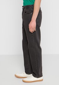 Pantalones de mezclilla negros con un diseño de pierna recta, estilo de cinco bolsillos y costuras visibles. Llevados con zapatillas blancas.