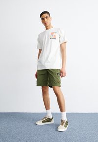 Men's cream t-shirt with colorful graphic pocket, paired with olive green shorts featuring zip pockets, and beige-green sneakers with white socks.