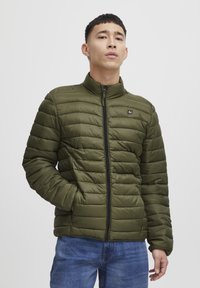 Olive green padded jacket with a stand-up collar, front zipper, and horizontal quilted pattern. Paired with blue jeans.