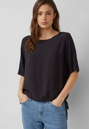 Kobieta z długimi brązowymi włosami, ubrana w luźną czarną bluzkę z krótkim rękawem i jasnoniebieskie dżinsy, z rękami w kieszeniach, stojąca na jednolitym tle.