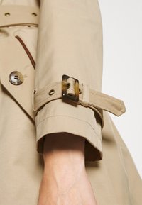 Rukáv béžového trenčkotového kabátu s přezkou ve tvaru želvoviny zapnutou nad zápěstím a viditelným knoflíkem poblíž lokte.