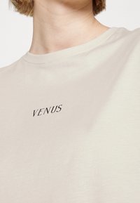 Camiseta de algodón beige claro con cuello redondo que presenta el texto negro "VENUS" en el pecho. Textura suave y ajuste casual.