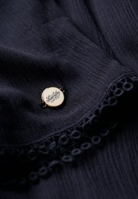 Marineblåt stof med en tekstureret overflade præsenterer et rundt, guldfarvet metaltag præget med "Superdry" og sorte tråd accenter.