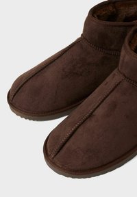 Smeđe suede papuče s zaobljenim vrhom i niskim dizajnom oko gležnja. Sadrže vidljive šavove duž šavova i teksturiranu potplat.