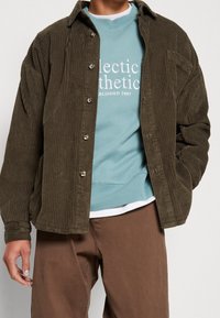 Olivgrünes Cordjackett mit lockerem Schnitt, ausgestattet mit silbernen Knöpfen und strukturiertem Stoff, darüber getragen ein hellblaues Sweatshirt.