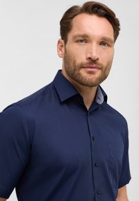 Camicetta blu navy a maniche corte con colletto strutturato, taschino frontale e dettagli a motivo all'interno del colletto; realizzata in tessuto liscio.