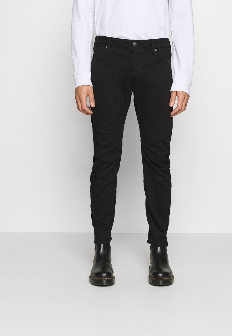Homme portant un pantalon noir slim associé à des bottes en cuir noires et une chemise blanche à manches longues, debout devant un fond uni.