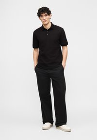 Νέος άνδρας που στέκεται με τα χέρια στις τσέπες, φορώντας μαύρο πόλο με κοντά μανίκια, μαύρο παντελόνι με φαρδιά μπατζάκια και λευκά αθλητικά παπούτσια σε απλό φόντο.