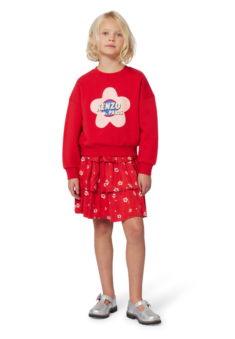 Felpa rossa con un logo a forma di fiore che presenta "KENZO PARIS", abbinata a una gonna a balze floreale rossa e scarpe con cinturino argentate.