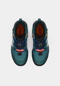 Sapatos de exterior em azul turquesa com um upper texturizado, apresentam uma sola de borracha preta, atacadores azuis e detalhes laranja, rotulados como Gore-Tex e com cordões elásticos.