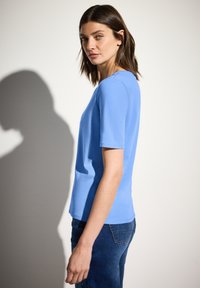 Světle modrý top s krátkým rukávem a kulatým výstřihem, vyrobený z měkkého materiálu, spárovaný s tmavě modrými džíny. Hladká textura s přiléhavým designem.