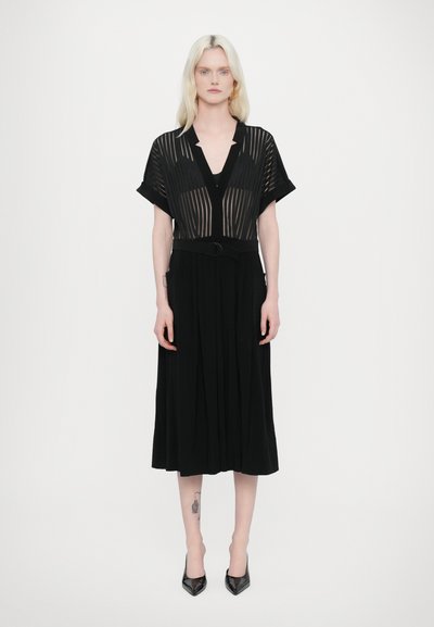 Robe noire avec un corsage en dentelle rayée et transparent, décolleté en V profond, manches courtes et taille ceinturée, descendant jusqu'à une jupe plissée.