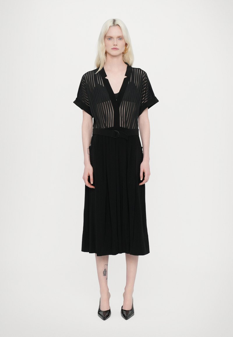 Vestido negro con un corsé de rayas transparente y un escote en V profundo, mangas cortas y cintura ceñida con un cinturón, que cae hacia una falda plisada.