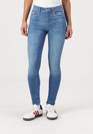 Vrouw met lichtblauwe skinny jeans en witte sneakers met rode strepen, staand tegen een effen witte achtergrond.