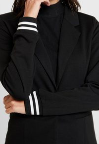 Blazer nero con una vestibilità sartoriale caratterizzato da strisce bianche sui polsini a coste e un top a collo alto sotto. Tessuto dalla texture liscia.