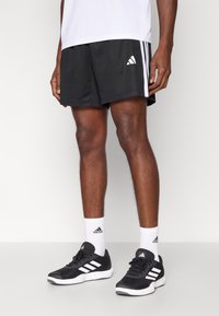 Pantalones cortos deportivos negros con rayas blancas y un logo de Adidas. Combinados con zapatos negros y calcetines tobilleros blancos con el logo de Adidas.