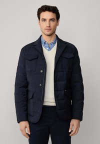 Marineblauwe pufferjack met meerdere voorzakken, knoopsluiting en een gladde textuur. Draag het over een lichte trui en een gestreept shirt.